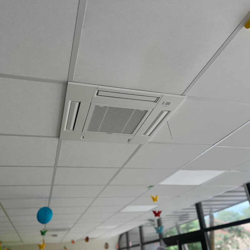 Climatiseur Cassette 4-voies : Intégration dans Faux Plafond. Climatiseur cassette Mitsubishi blanc encastré dans un faux plafond modulaire au-dessus de décorations colorées (papillons et boules).
