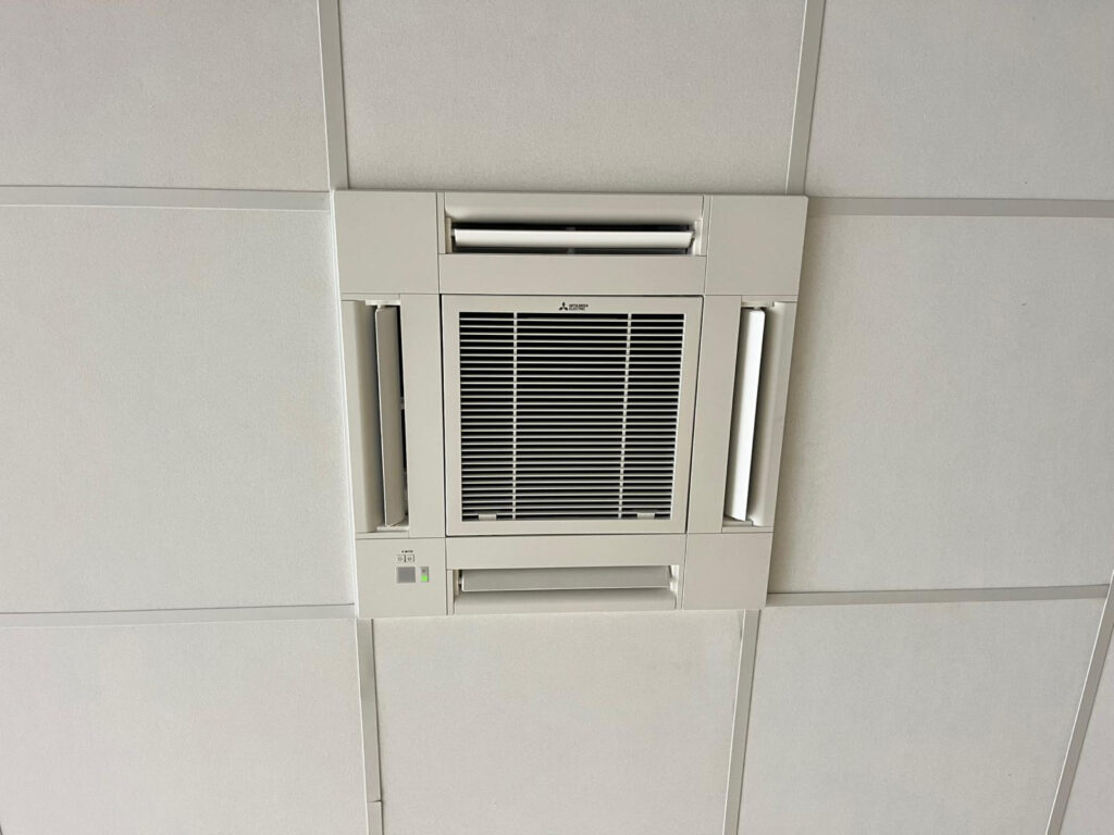 Climatiseur cassette Mitsubishi pour faux plafond Climatiseur cassette Mitsubishi Electric 4-voies encastré dans un faux plafond. Grille centrale et volets de ventilation latéraux ouverts.