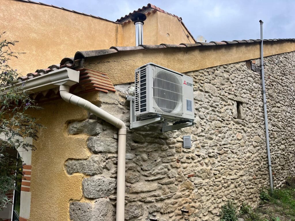 Climatiseur Mitsubishi : intégration réussie sur mur de pierre. Climatiseur Mitsubishi Electric sur console contre une façade en pierres apparentes. Une gouttière beige descend le long du mur et du crépi ocre.