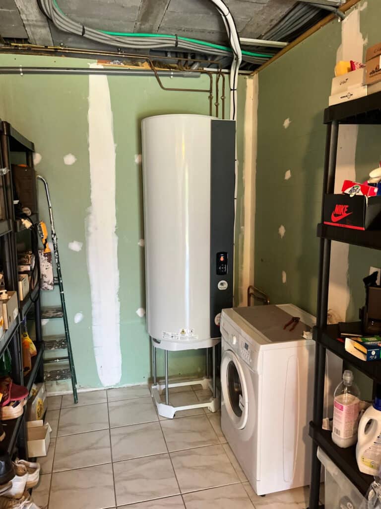 Chauffe-eau moderne : installation en buanderie/garage en rénovation. Buanderie/garage en rénovation. Chauffe-eau moderne blanc sur pied, machine à laver, étagères de rangement, murs inachevés et tuyauteries.
