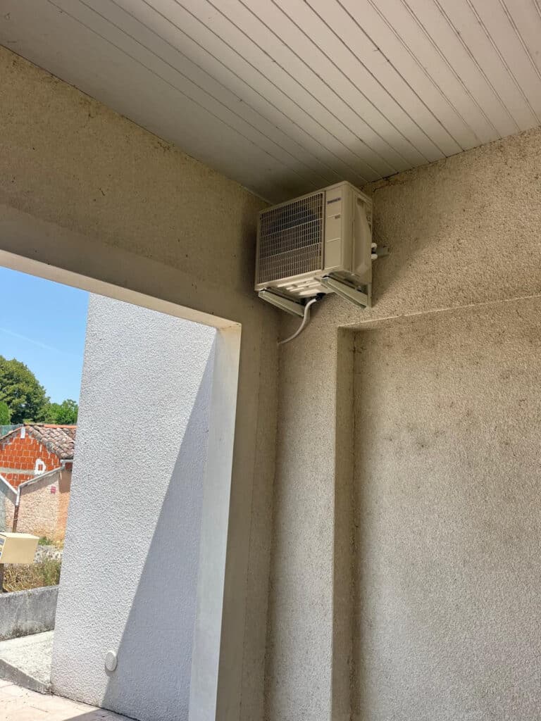 Climatiseur exterieur Mitsubishi : installation murale reussie Unité extérieure de climatiseur Mitsubishi (R32) installée en hauteur sur un mur crépi sous un porche. Vue sur des tuiles.
