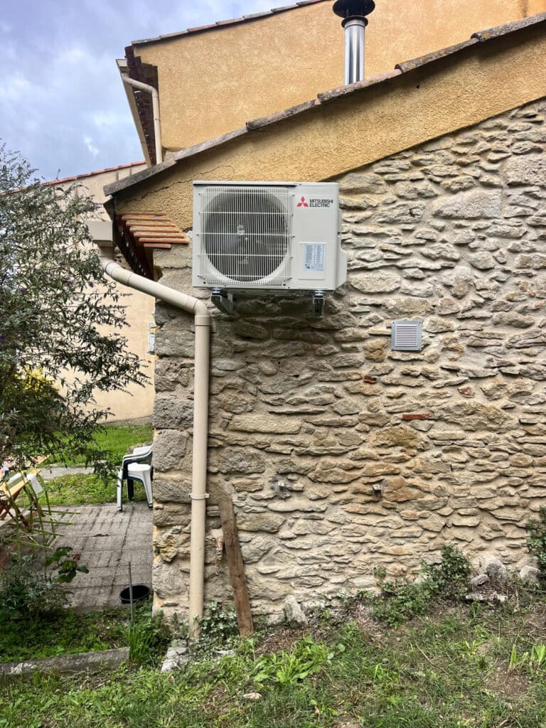 Climatisation Mitsubishi Electric : pose sur mur pierre. Unité extérieure Mitsubishi Electric fixée solidement sur un mur en pierres apparentes. Gouttière beige et ventilation de façade.