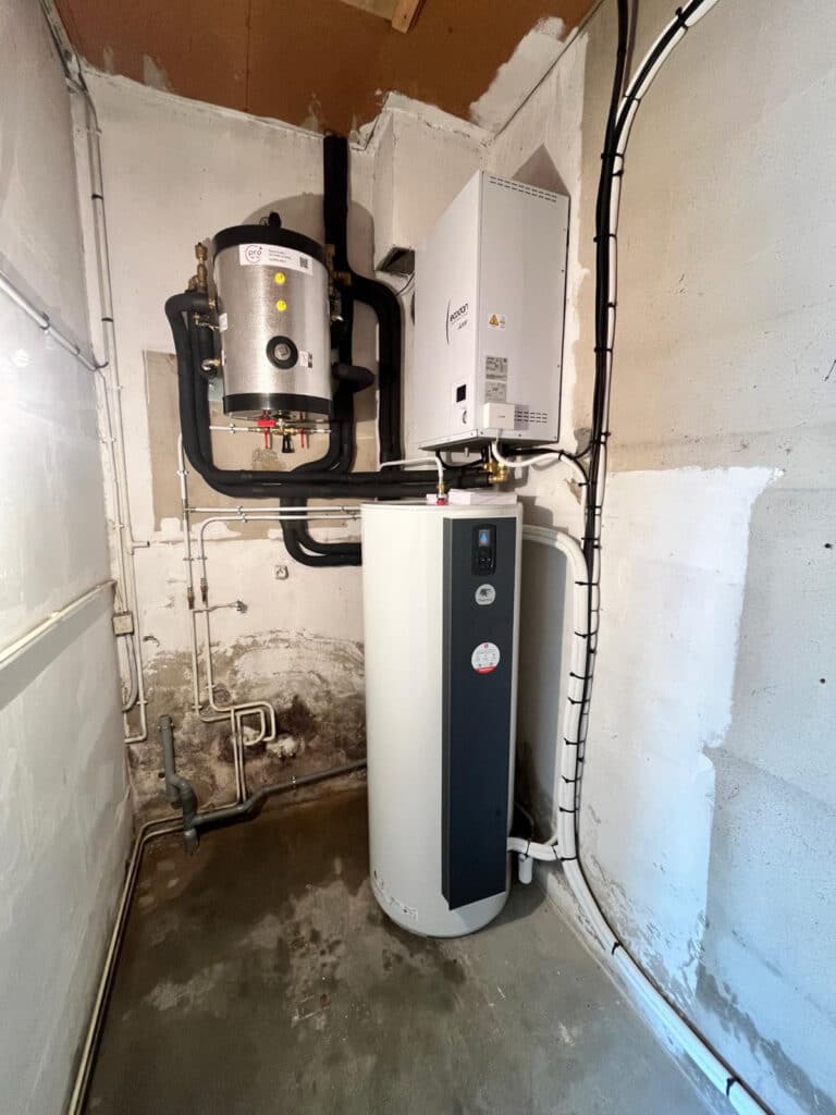 Pompe à chaleur Ecodan : installation professionnelle en sous-sol. Système de pompe à chaleur Mitsubishi Ecodan (unité murale) et son ballon tampon vertical, installé dans un local technique brut. Tuyauterie isolée noire.