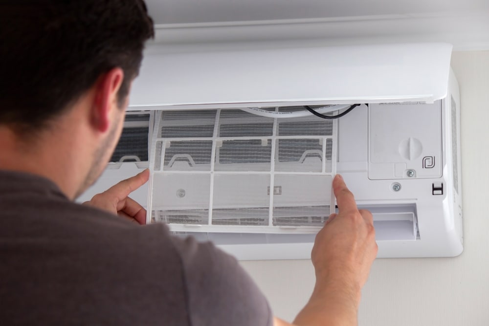 Climatiseur : comment retirer et nettoyer le filtre ? Gros plan d'un homme retirant le filtre poussiéreux de l'unité intérieure d'un climatiseur mural pour entretien.